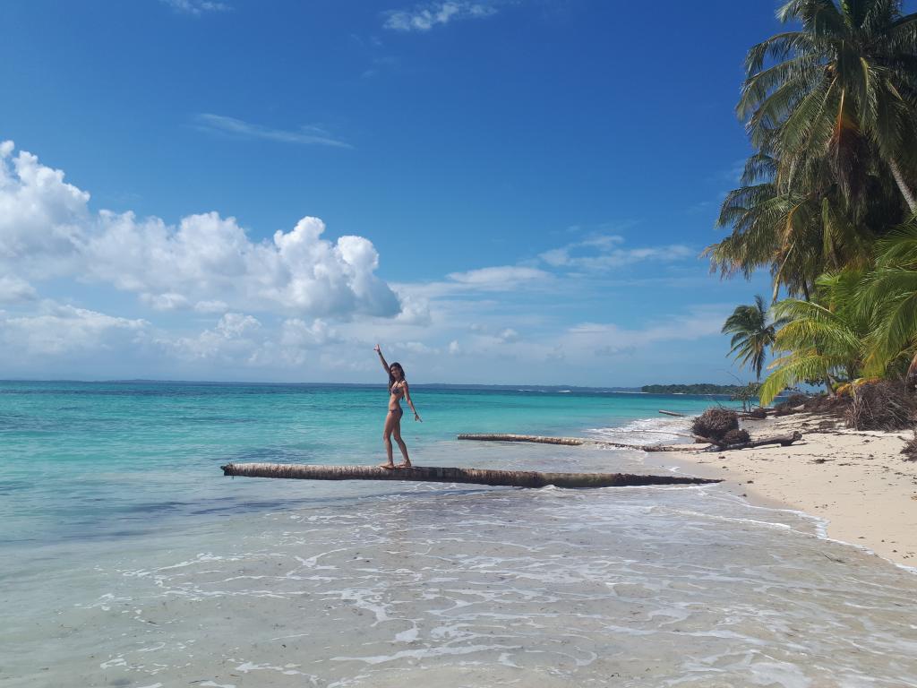 Chica caminando encima de un gran tronco que se encuentra en la orilla de una playa paradisíaca de arena blanca y aguas turquesas, custodiada por palmeras.
Un paraíso tropical que todo el mundo debería incluir al viajar a Panamá