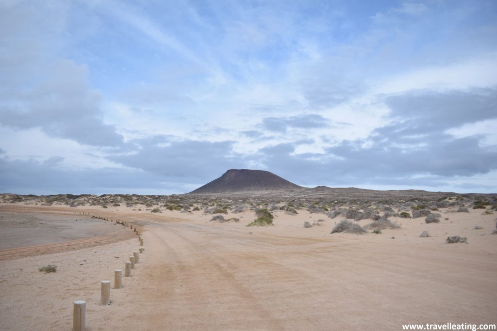 Camino de arena junto a la playa tras el cual se ven dunas y, más al fondo, un volcán.