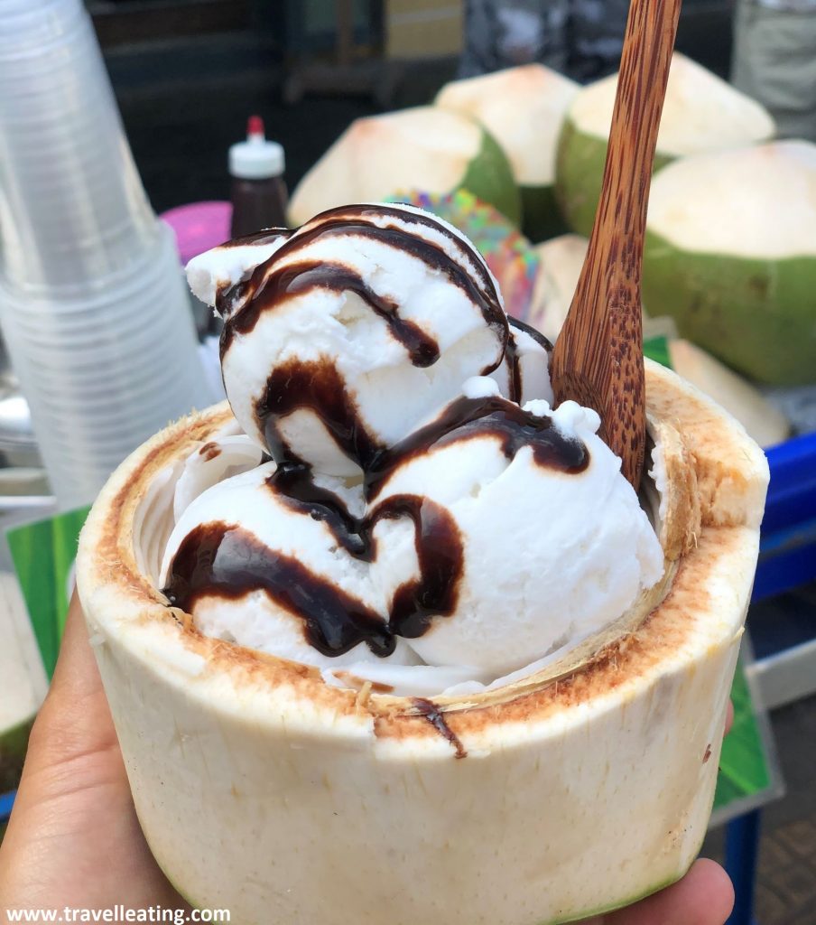 Tres bolas de helado de coco servidas en un coco fresco que hace de recipiente. Tiene chocolate negro por encima y una cuchera de bambú clavada.