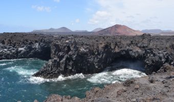 Tramo de costa formado por riscos y acantilados de roca volcánica repletos de cavidades y cuevas, tras el cual se observa un paisaje volcánico.