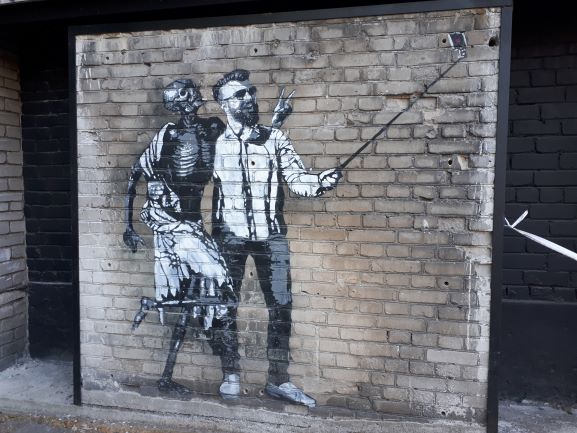 Mural de arte callejero en el que se ve un chico joven, de estilo hípster, haciéndose un selfie con el movil con un esqueleto.