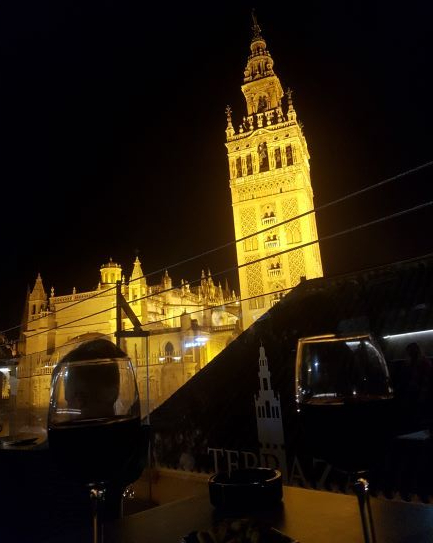 Vistas de la Giralda iluminada de noche desde una mesa en una terraza. Sobre la mesa podemos ver dos copas de vino tinto.
