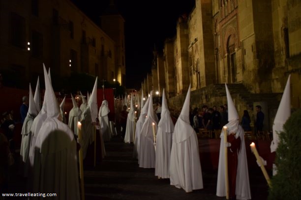 Procesión nocturna frente a la Mezquita-Catedral de Córdoba. Se ve un grupo de personas con túnicas y capuchas blancas, las cuales aguantan un cirio encendido.
