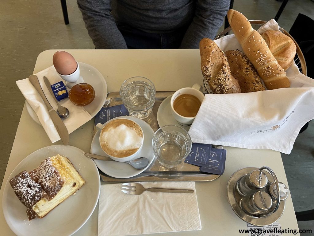 Mesa servida con un desayuno generoso vista desde arriba. Se ven dos cafés (uno con crema y el otro solo), una strudel de queso, una cesta con 4 panes diferentes y un recipiente con un huevo duro, mantequilla y mermelada.