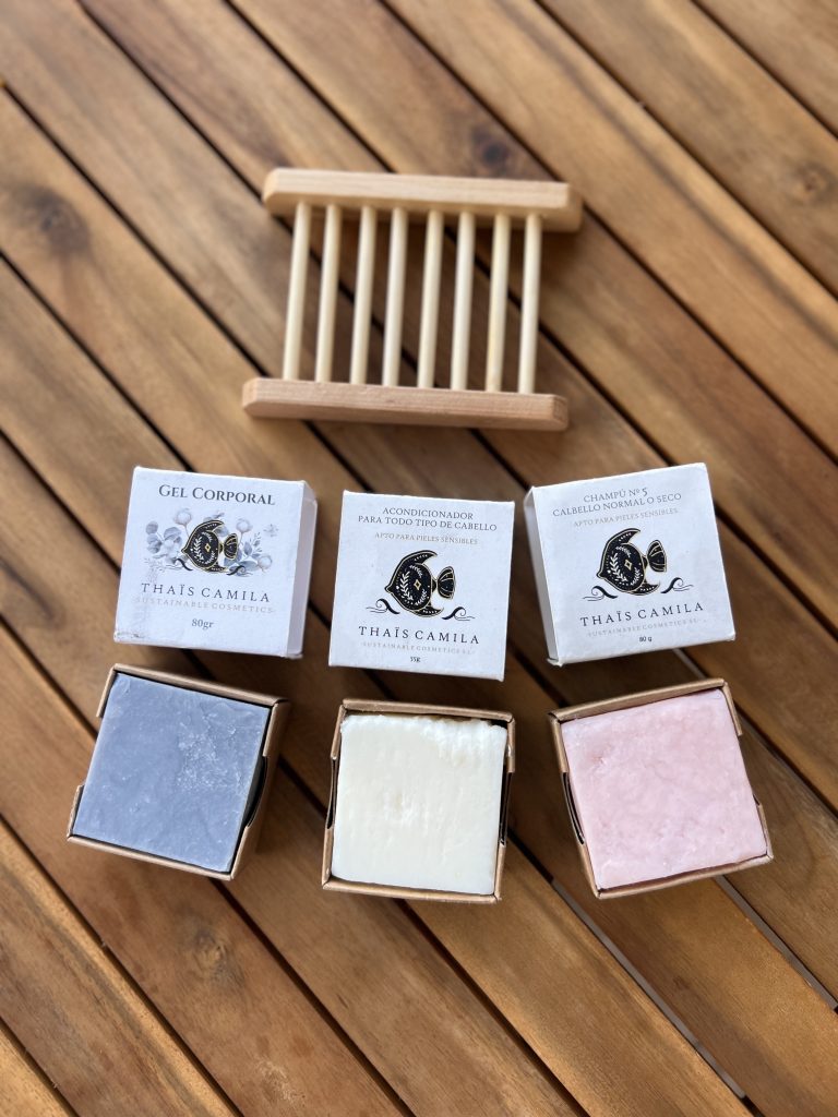 Pastillas solidas de champú, acondicionador y jabón de cuerpo (de color azul, blanco y rosa) encima de una mesa de madera y junto a sus cajas y a una jabonera de bambú