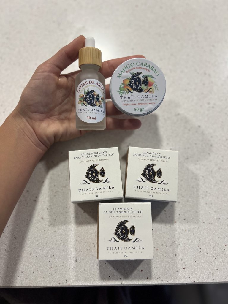 tres cajas que contienen jabón, champú y acondicionadores sólidos, junto a una mano que sujeta un bote de cristal con aceite de argán y un bote de manteca de karité.