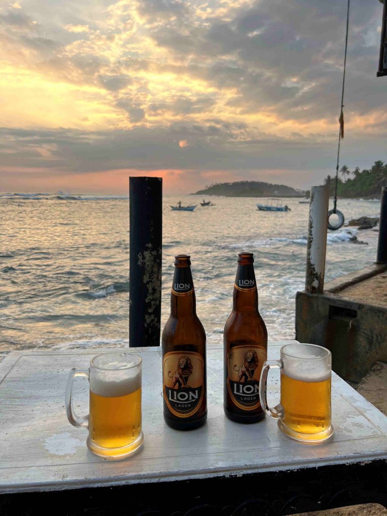 Cervezas Lion servidas en un chiringuito junto al mar, mientras los pescadores tradicionales pescan en el mar al atardecer.